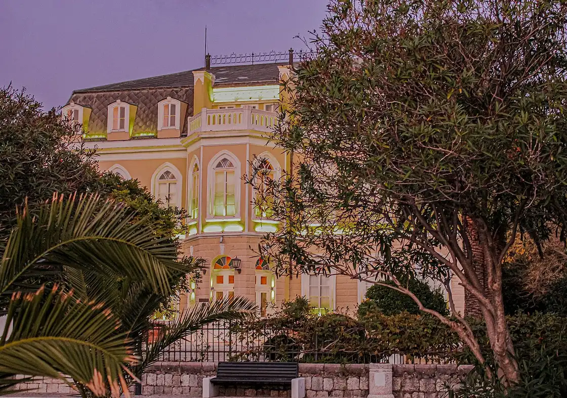 King Nikola's Palace in Bar floodlit at dusk behind its garden fence and trees