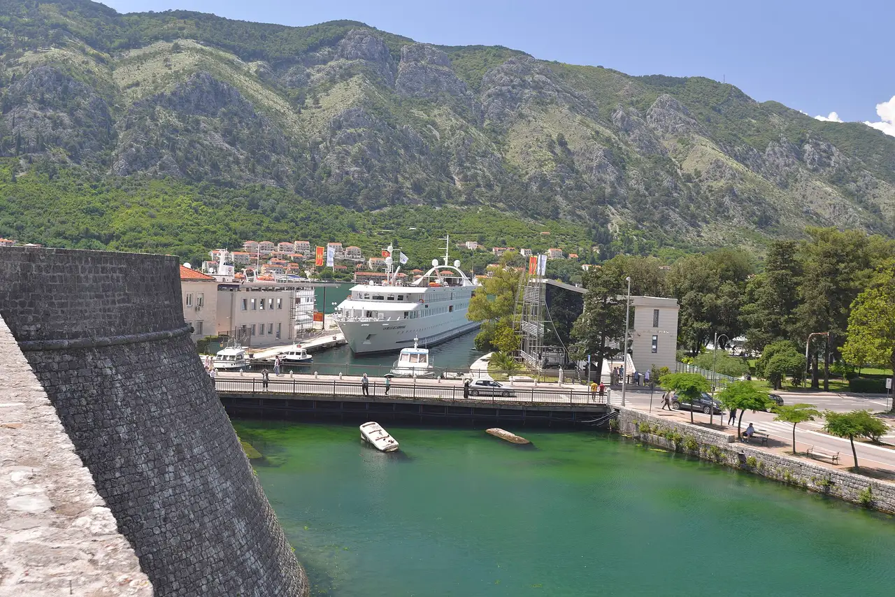 Boats at the Kotor harbour quay below the old town walls, with a cruise ship moored behind