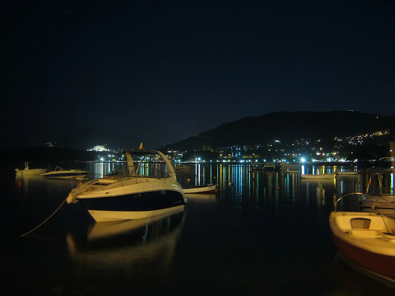 Boats moored off Bečići after dark, the resort strip lit along the bay behind them