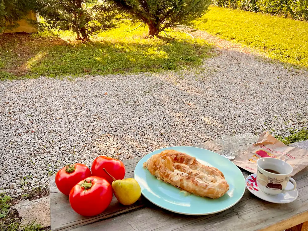 A burek pastry on a plate with tomatoes, a pear and a cup of coffee on an outdoor table