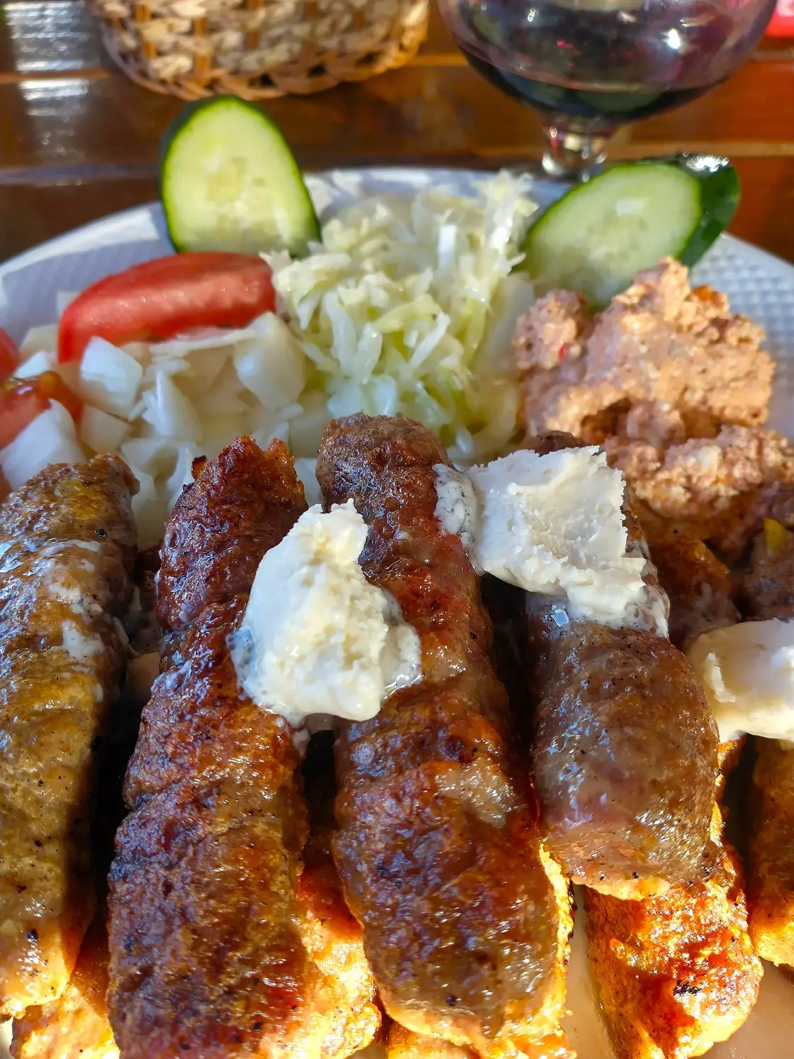 A plate of grilled cevapi with kajmak cream, shredded cabbage salad, tomato and cucumber