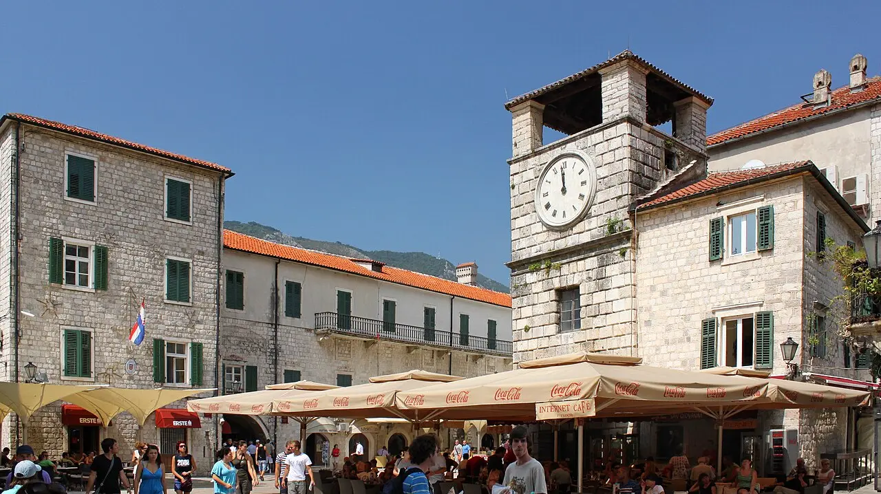Café and restaurant terraces packed under parasols on Kotor's Square of Arms, with the clock tower behind