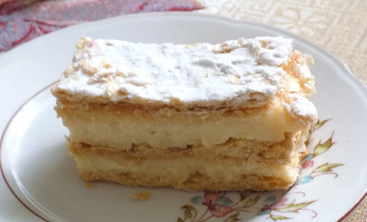A slice of Kotorska krempita custard cake dusted with icing sugar on a plate
