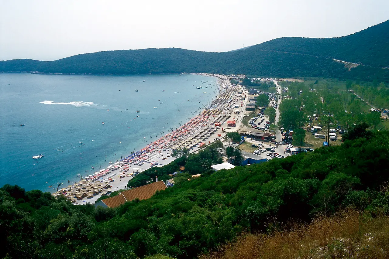 Aerial view of Jaz beach: a long curved bay with rows of umbrellas, a campground and green hills behind