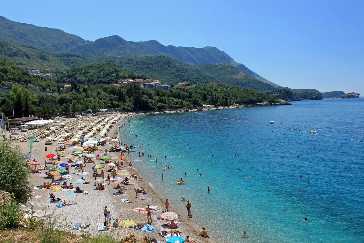 Kamenovo beach: turquoise water, a pebble beach lined with loungers and green mountains, with Sveti Stefan faint in the distance