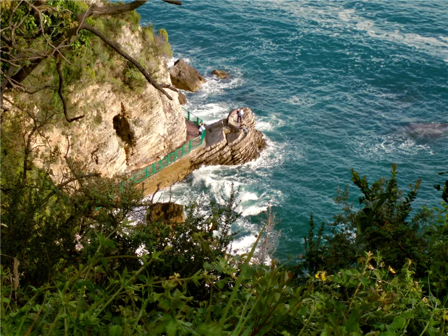 The cliff-side walkway with green railings leading around the rocks to Mogren beach, Budva