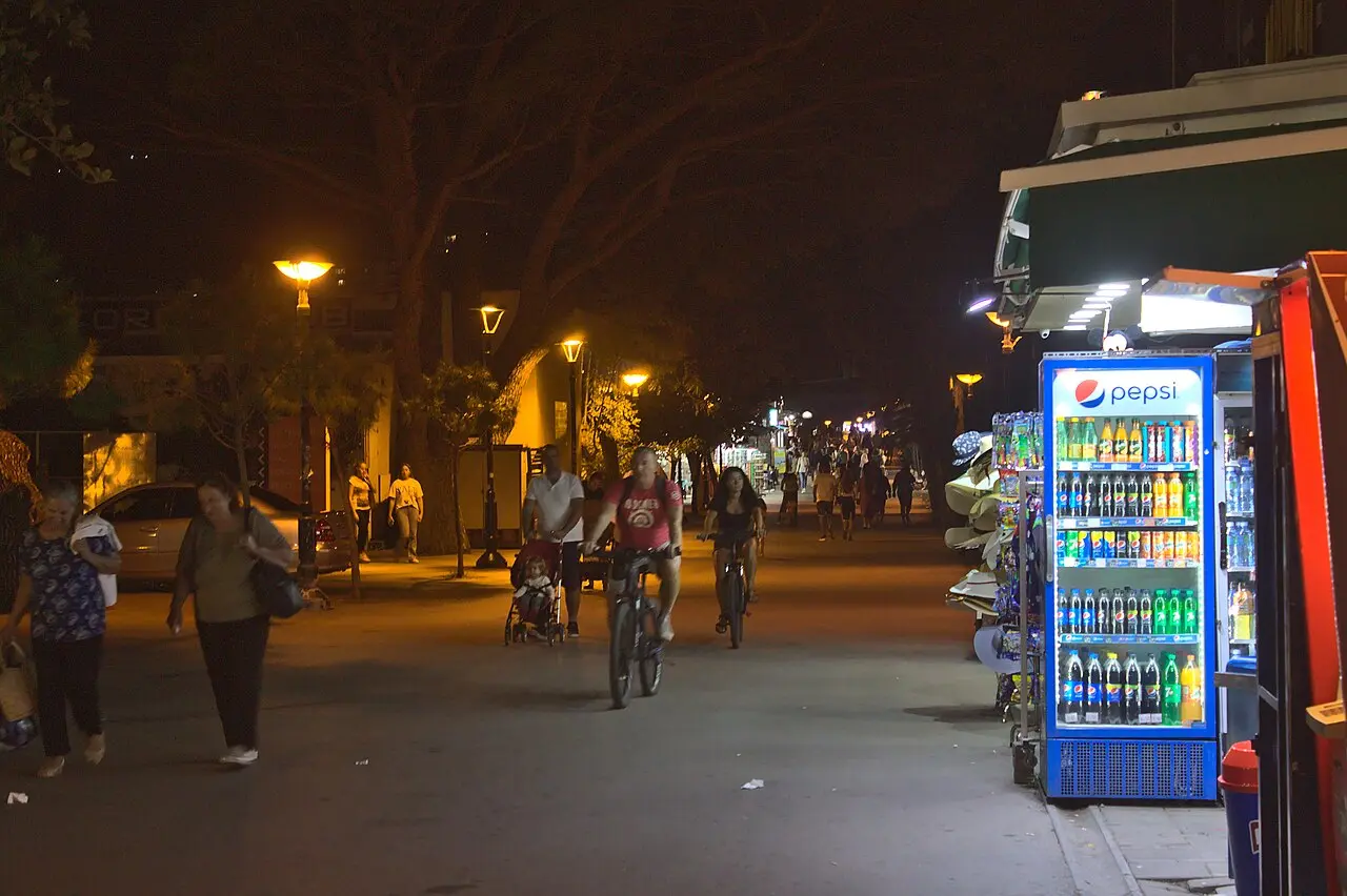 People walking and cycling along Budva's tree-lined seafront promenade at night past lit kiosks