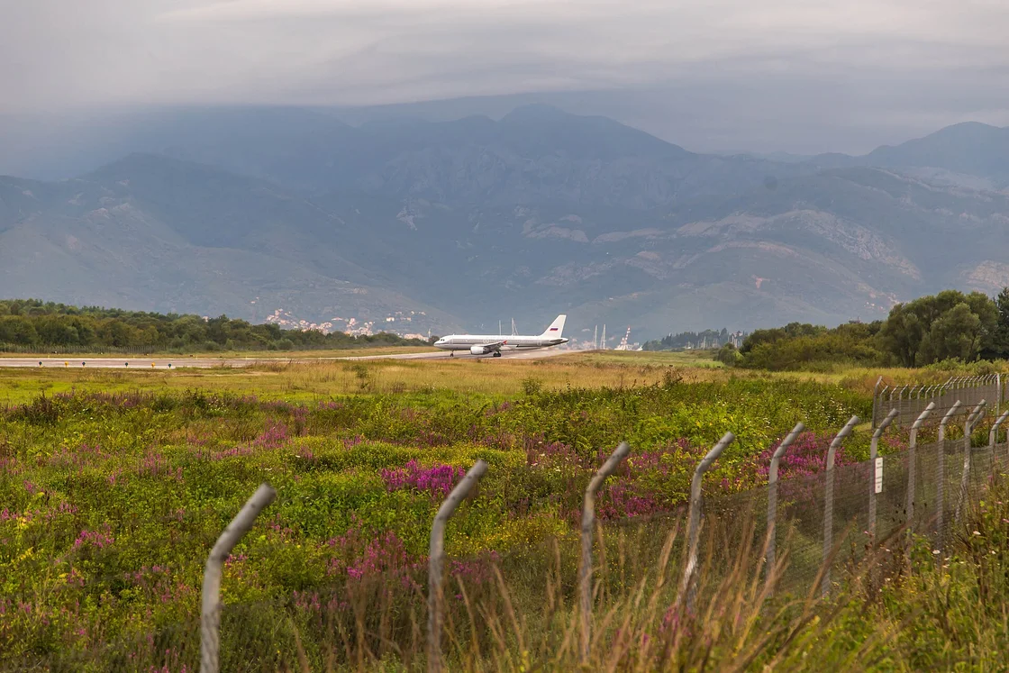 An airliner on the single runway of Tivat Airport with mountains behind, seen past the perimeter fence