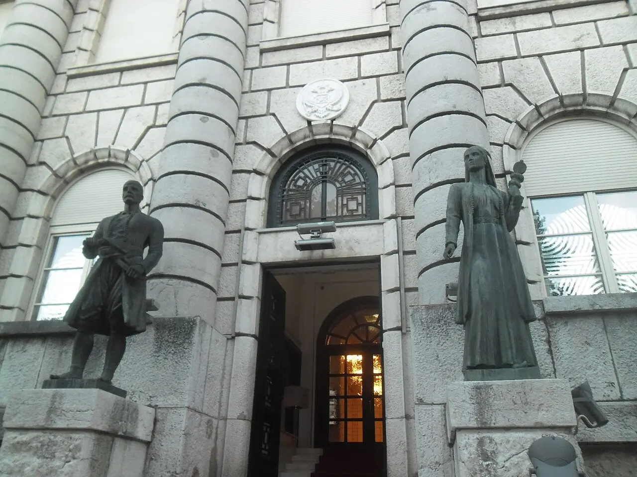 The stone entrance of a grand civic building in Cetinje flanked by two bronze statues