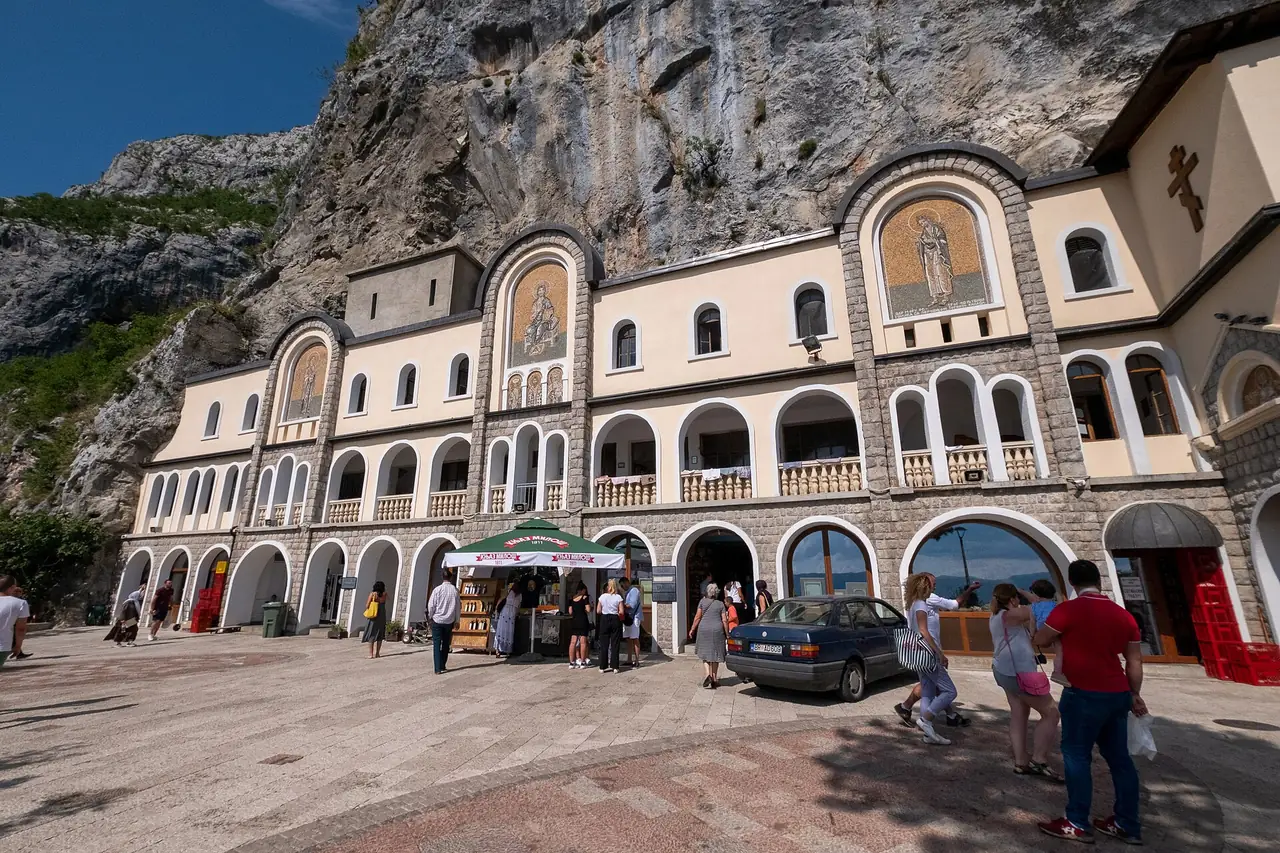 Lower Ostrog courtyard: arched mosaics, a paved square, a Knjaz Milos umbrella and a parked car