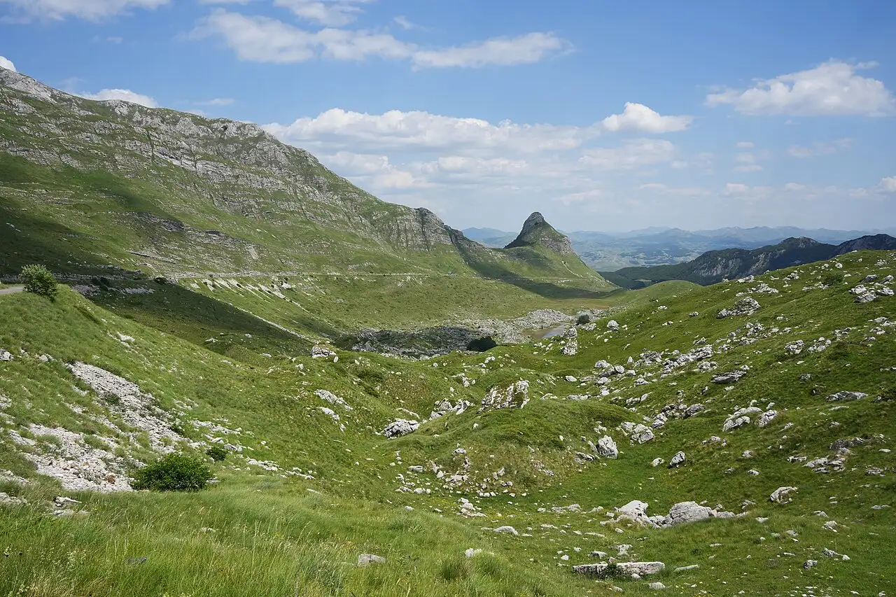 The green karst valley and lone rock peak near the Sedlo pass, the mountain road visible along the slope