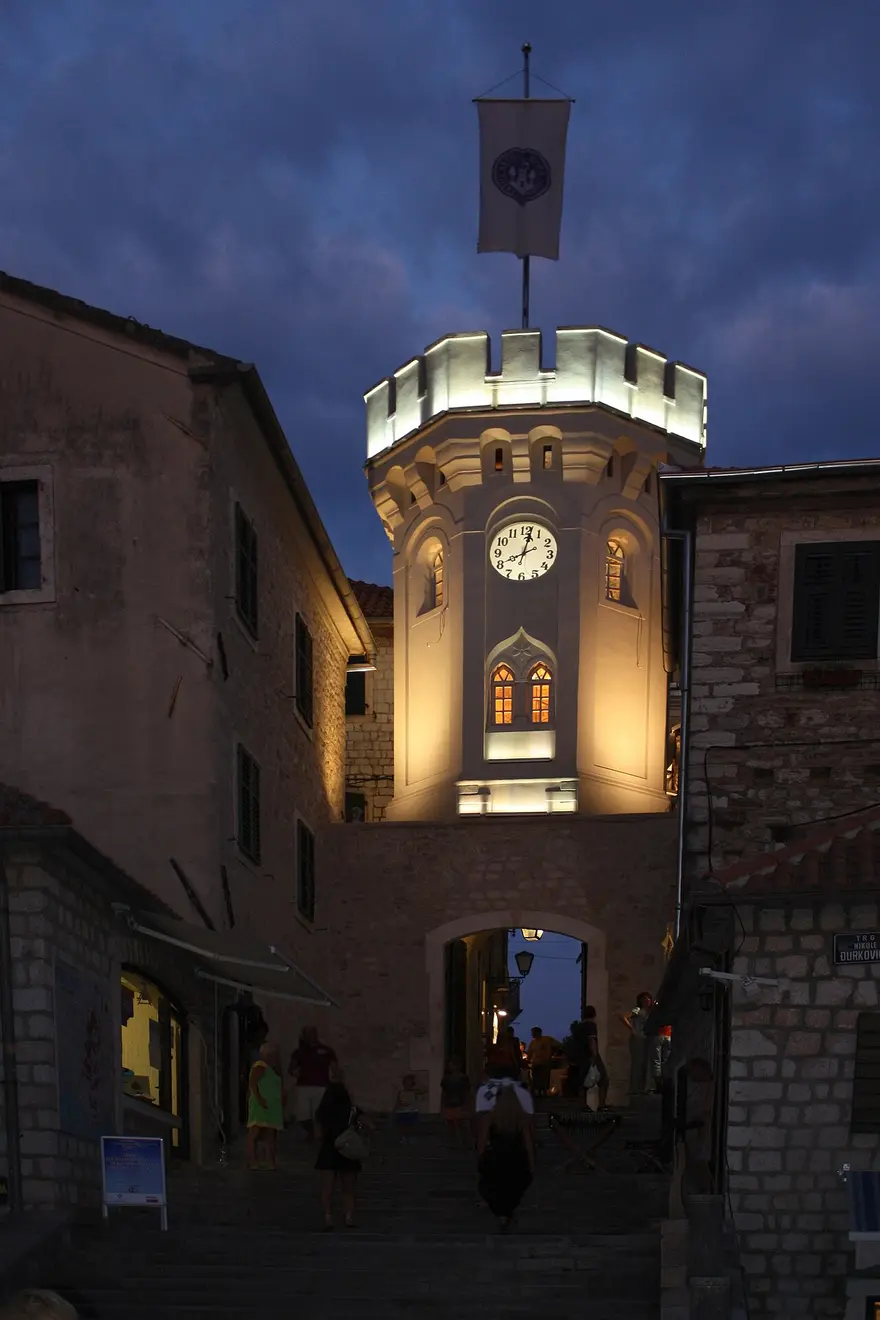 The illuminated Sahat Kula clock tower and its arched gate at the top of a stepped street in Herceg Novi at dusk