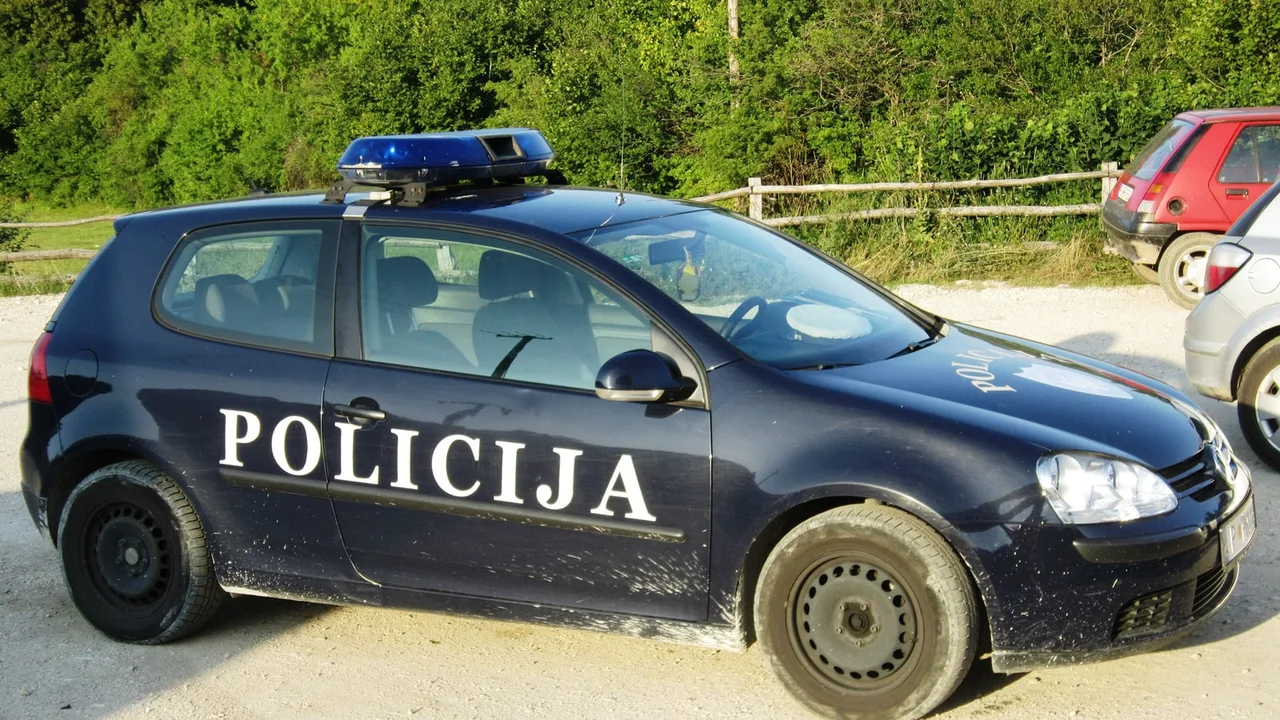 A dark blue Montenegrin police car marked POLICIJA, a Volkswagen Golf with a blue light bar, parked on a gravel lot beside a wooden fence