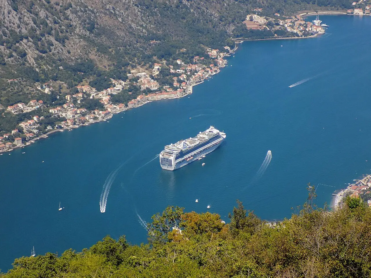 A large cruise ship moored in the narrow Bay of Kotor, dwarfing the waterfront town beneath the mountains