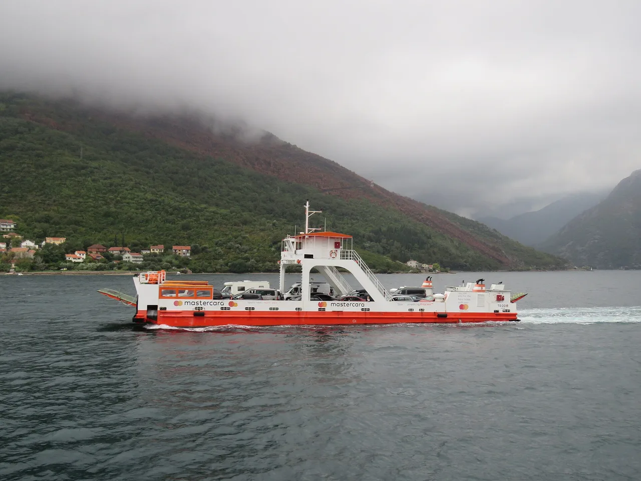 A car ferry loaded with cars and a campervan crossing the Bay of Kotor with mountains behind