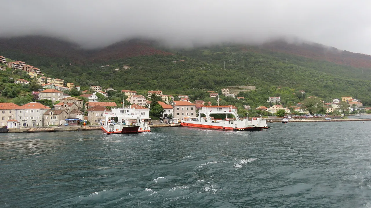 Two red and white car ferries at the pier in Lepetane village on the Bay of Kotor, low cloud on the hills behind