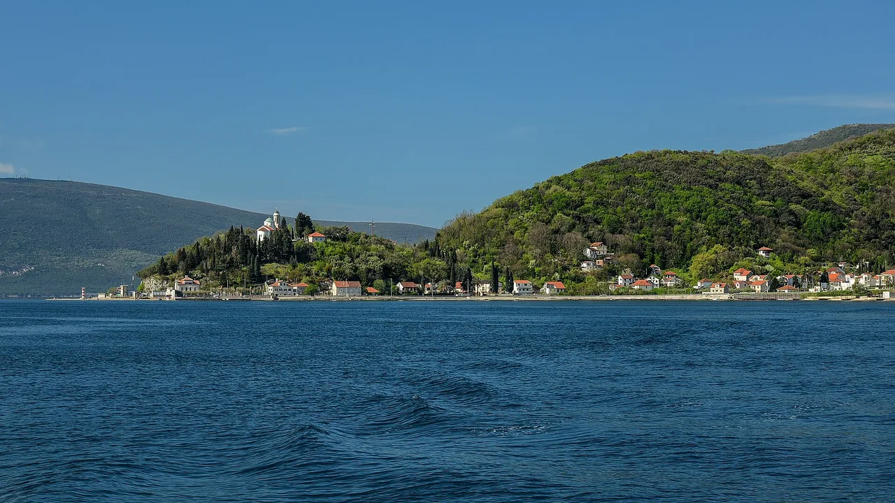 View across the Verige narrows of the Bay of Kotor towards the wooded headland and village on the far shore under a blue sky
