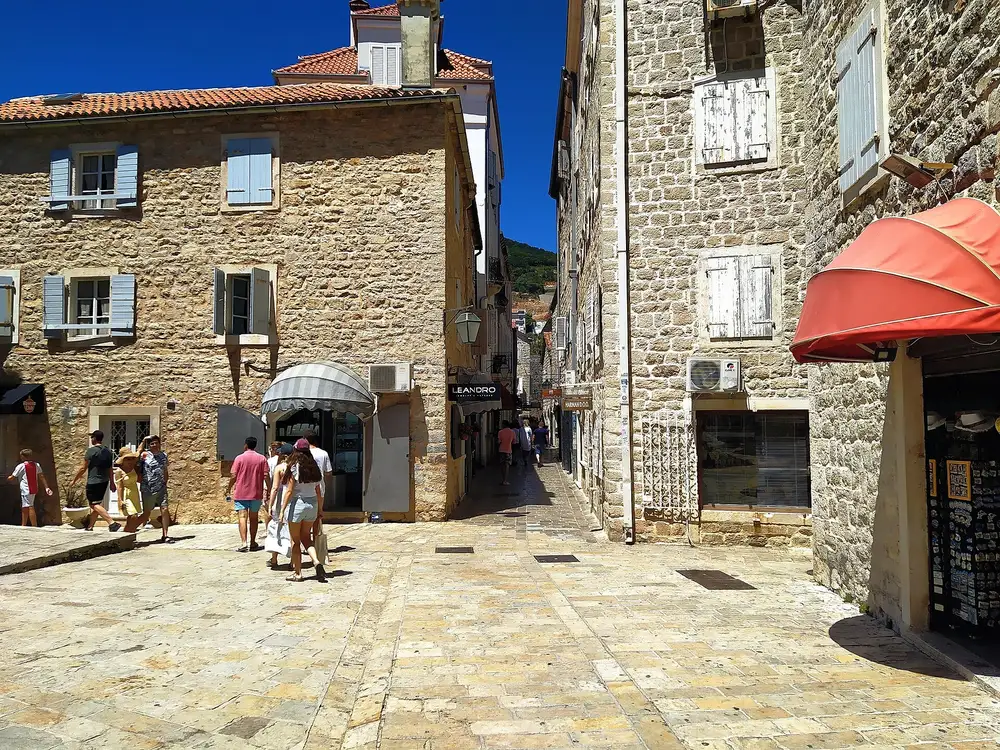 A sunny stone-paved lane in Budva Old Town lined with shops and stone houses, with people walking