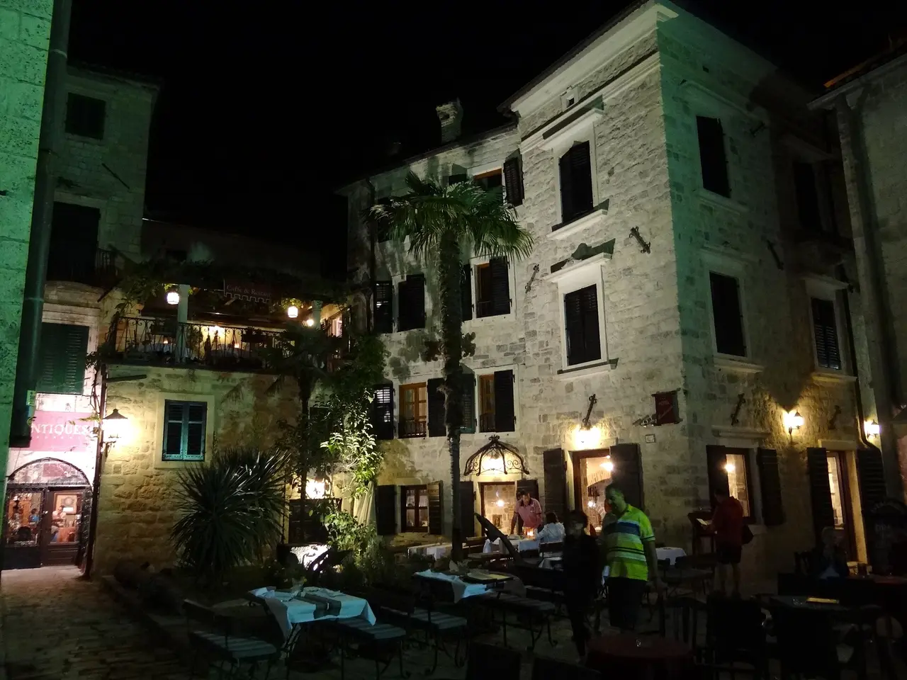 Diners at candlelit tables in a stone square in Kotor Old Town at night, lit by lanterns