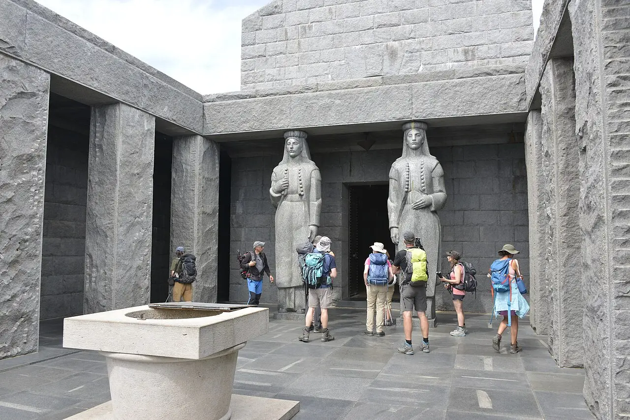 Two granite caryatid statues in Montenegrin folk dress flanking the entrance of the Njegoš Mausoleum, with visitors for scale