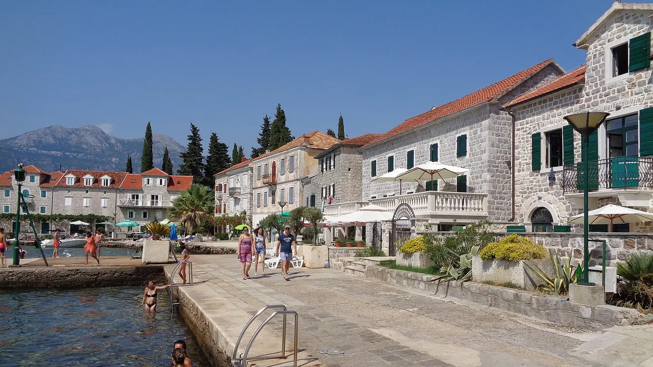 The stone waterfront of Rose village on the Lustica peninsula, with swimmers off the quay and mountains behind