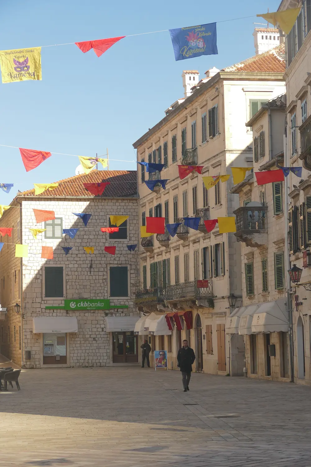 Carnival bunting and flags strung above an almost empty stone square in Kotor old town in February, one man walking