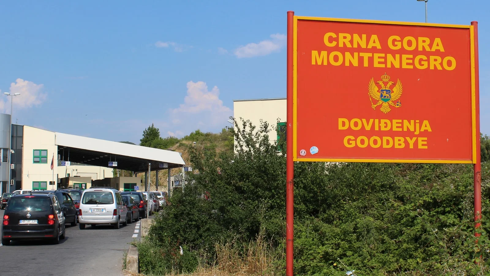 Cars queuing toward the control booths at a Montenegro-Albania border crossing, beside a red sign reading Crna Gora, Montenegro, Doviđenja, Goodbye
