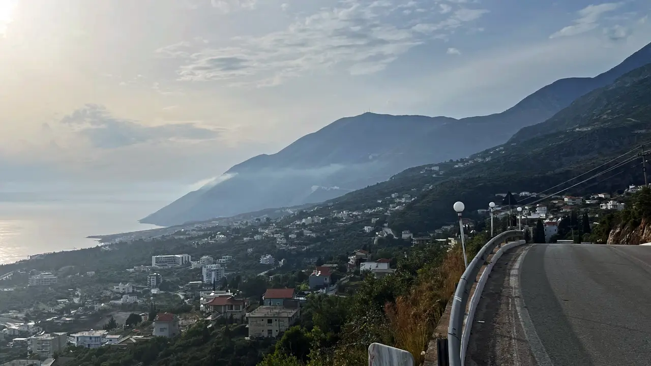 The Albanian Riviera coast near Dhermi, a winding road above scattered white houses with steep mountains dropping to the Ionian sea