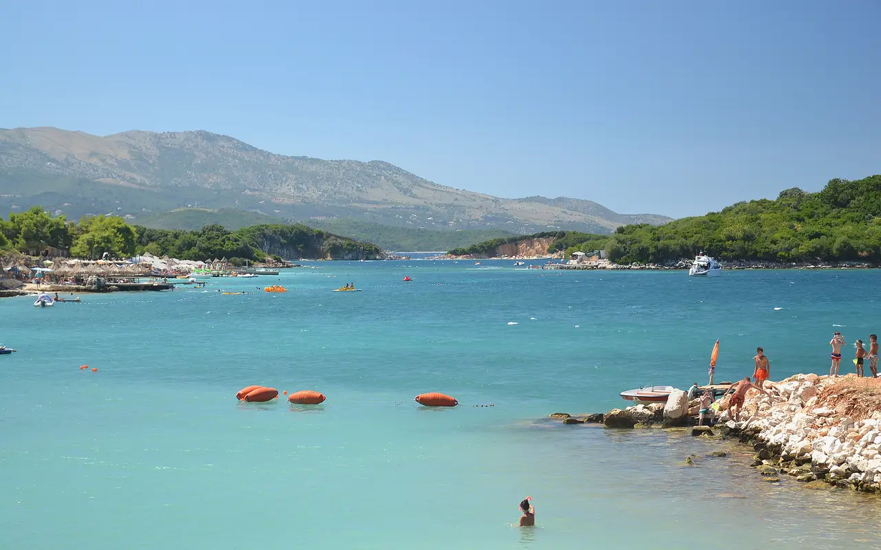 Swimmers and small boats in the bright turquoise shallows at Ksamil, with a wooded islet and mountains behind