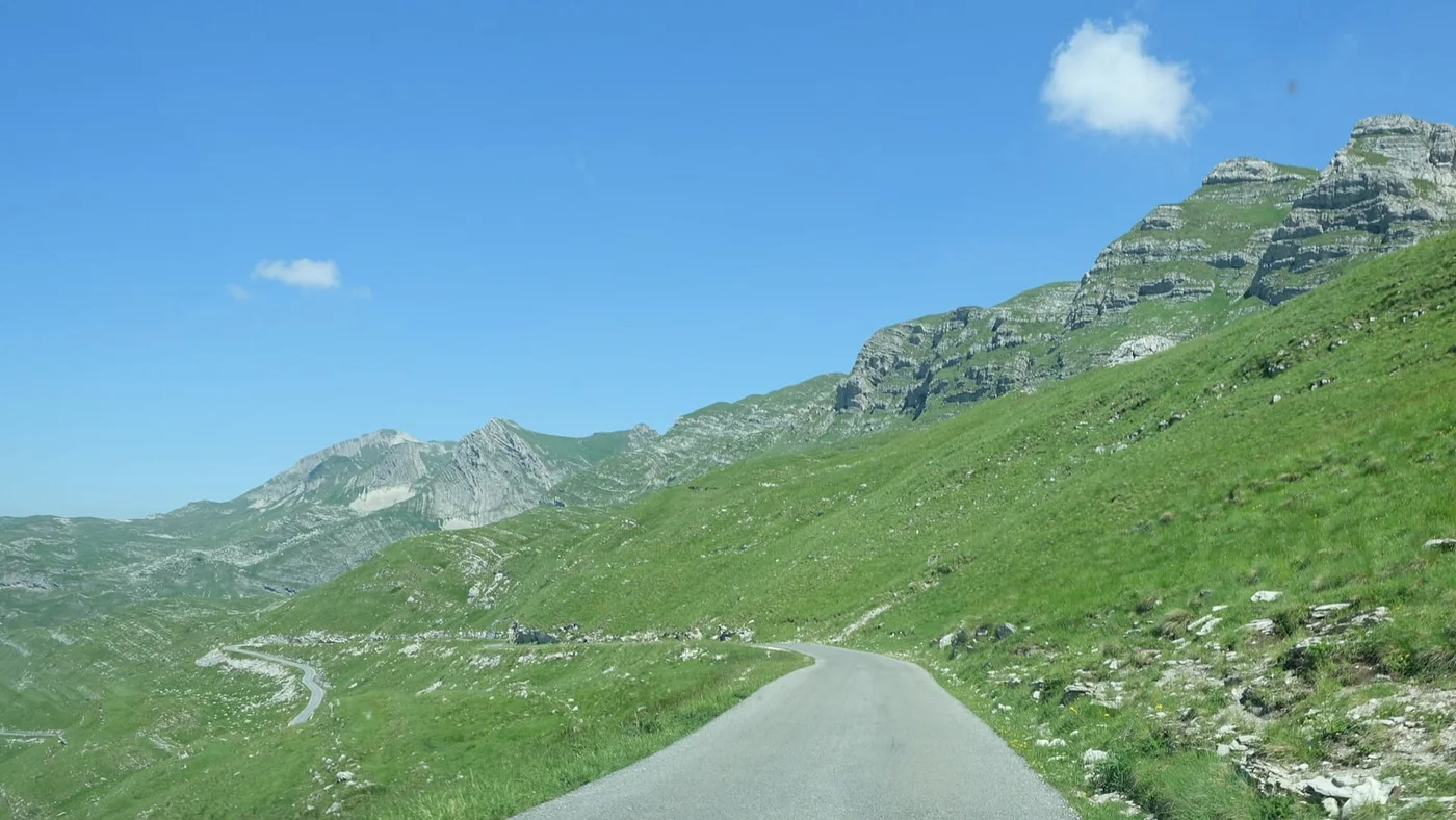 A narrow paved road crossing green alpine slopes below limestone peaks on the Sedlo pass in northern Montenegro