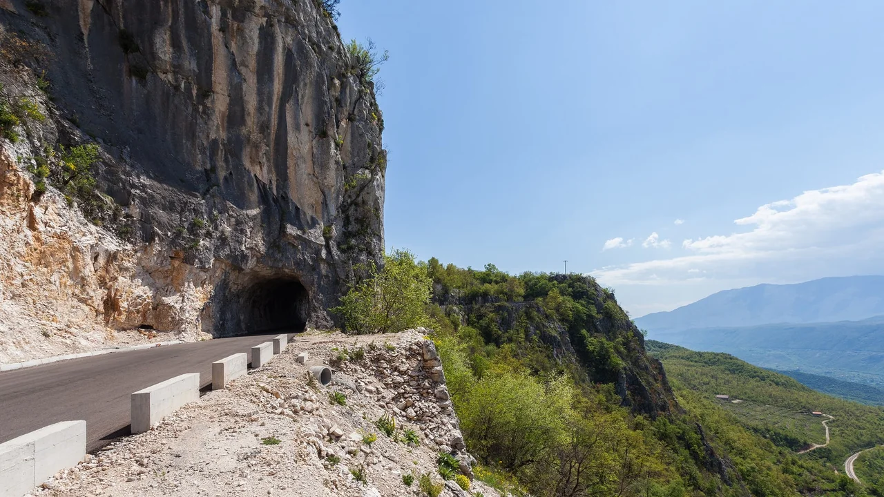 The road to Ostrog cut into the limestone cliff, with a rock tunnel and the Bjelopavlici valley below