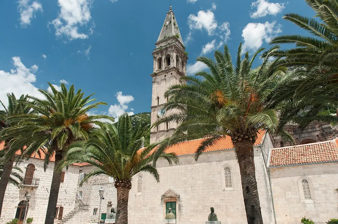 The tall stone bell tower of St Nicholas Church rising behind palm trees on the square in Perast