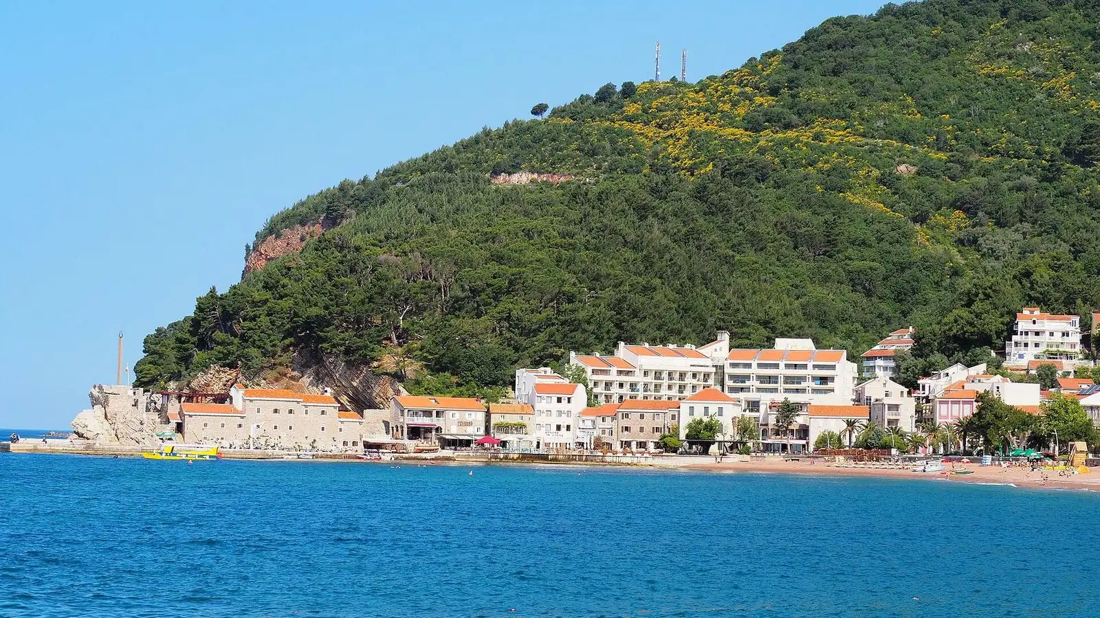 The town of Petrovac seen from the water, its red roofs strung along the beach under a green wooded headland, Montenegro