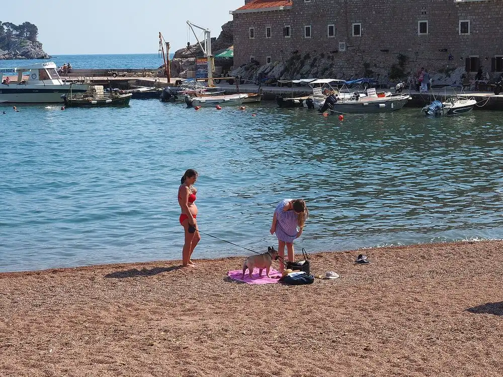 Small boats moored in the harbour of Petrovac beneath the stone wall of the old fortress, with children on the sand