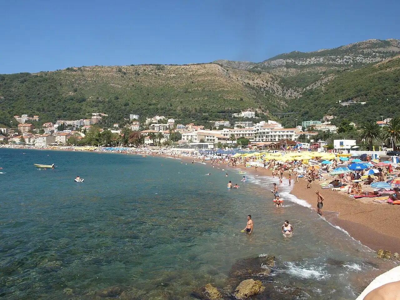 The reddish sand of Petrovac town beach with swimmers in the shallow water, seen from above