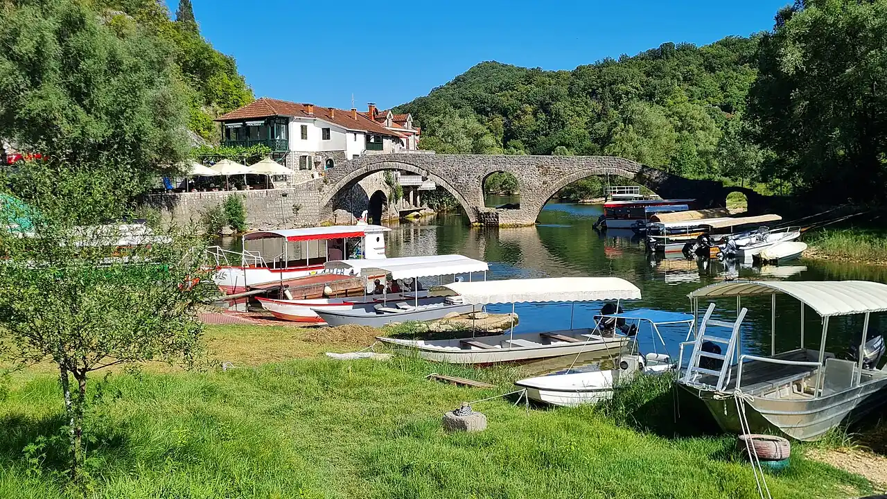 Danilo's double-arched stone bridge over the Crnojević River at Rijeka Crnojevića, with traditional wooden tour boats moored below and a riverside restaurant