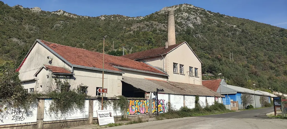An old disused fish-processing factory building with a tall brick chimney at Rijeka Crnojevića, now marking the start of boat tours