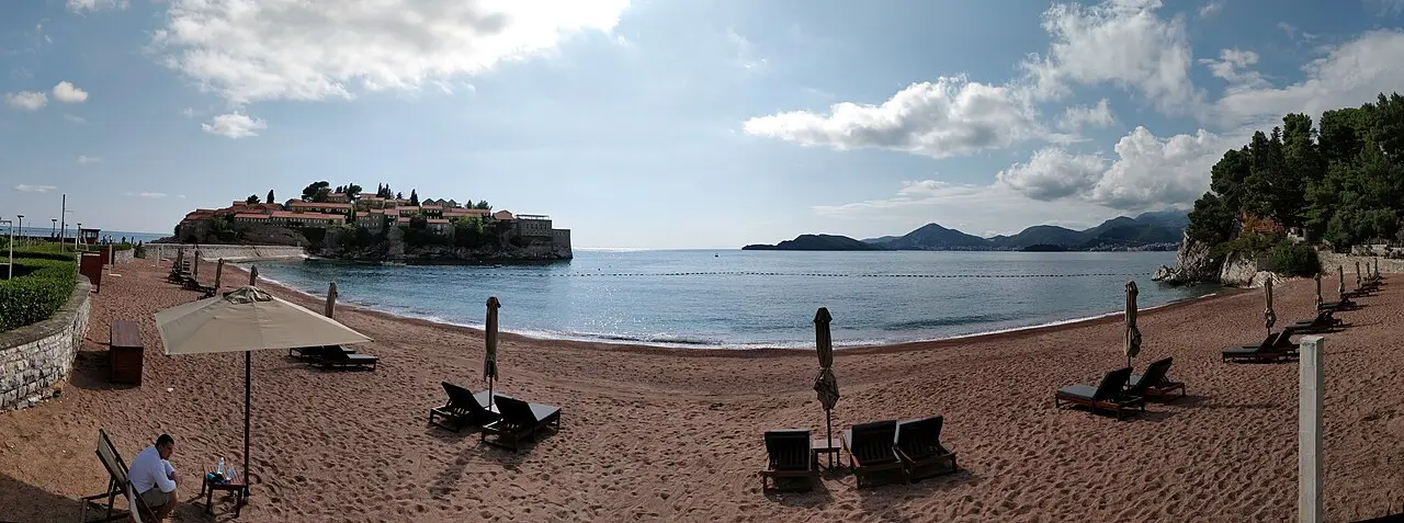 A wide panorama of the pink-sand beach and loungers below Sveti Stefan island, Montenegro