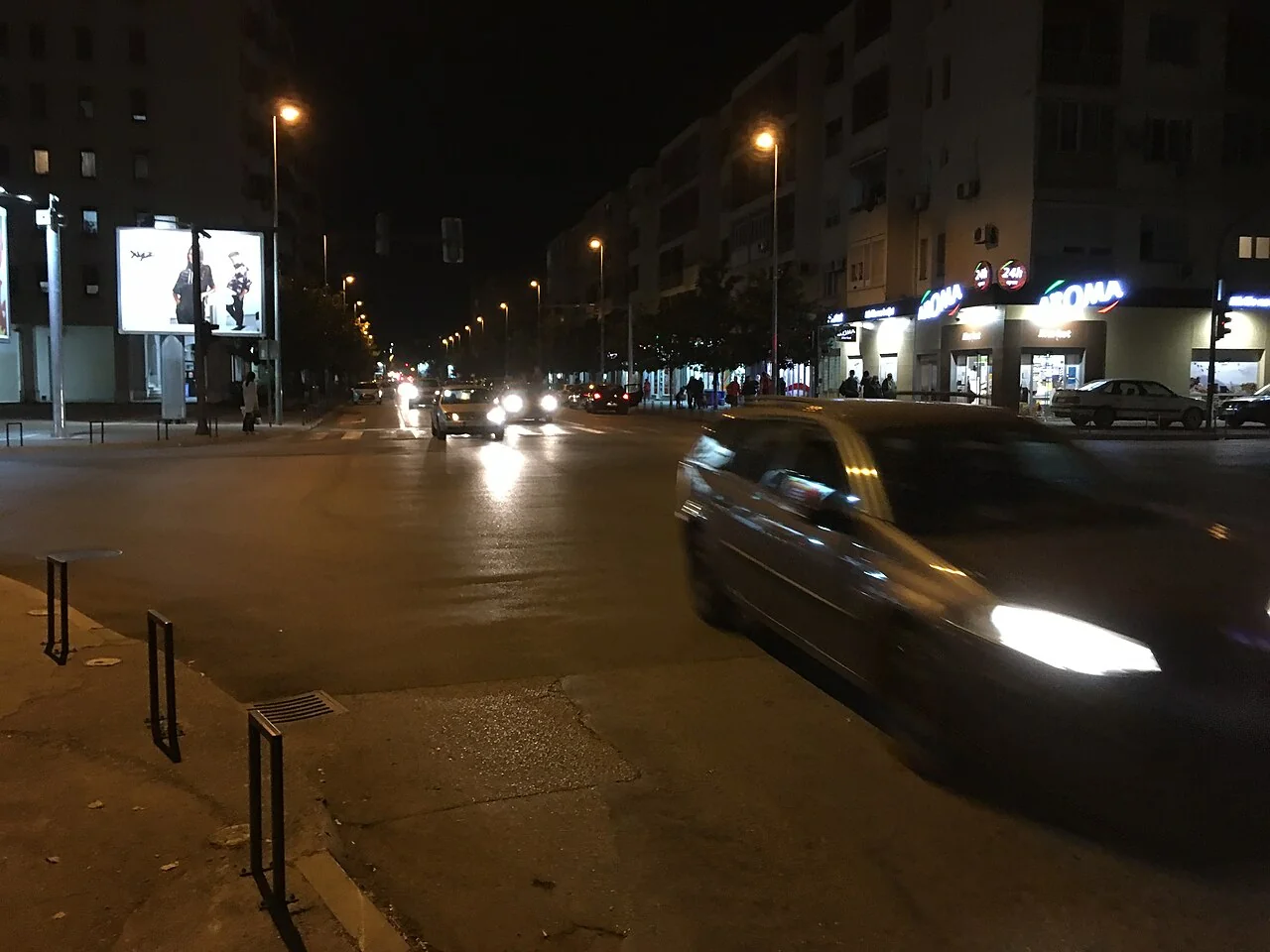 A car with its headlights on driving through a lit street in Podgorica at night