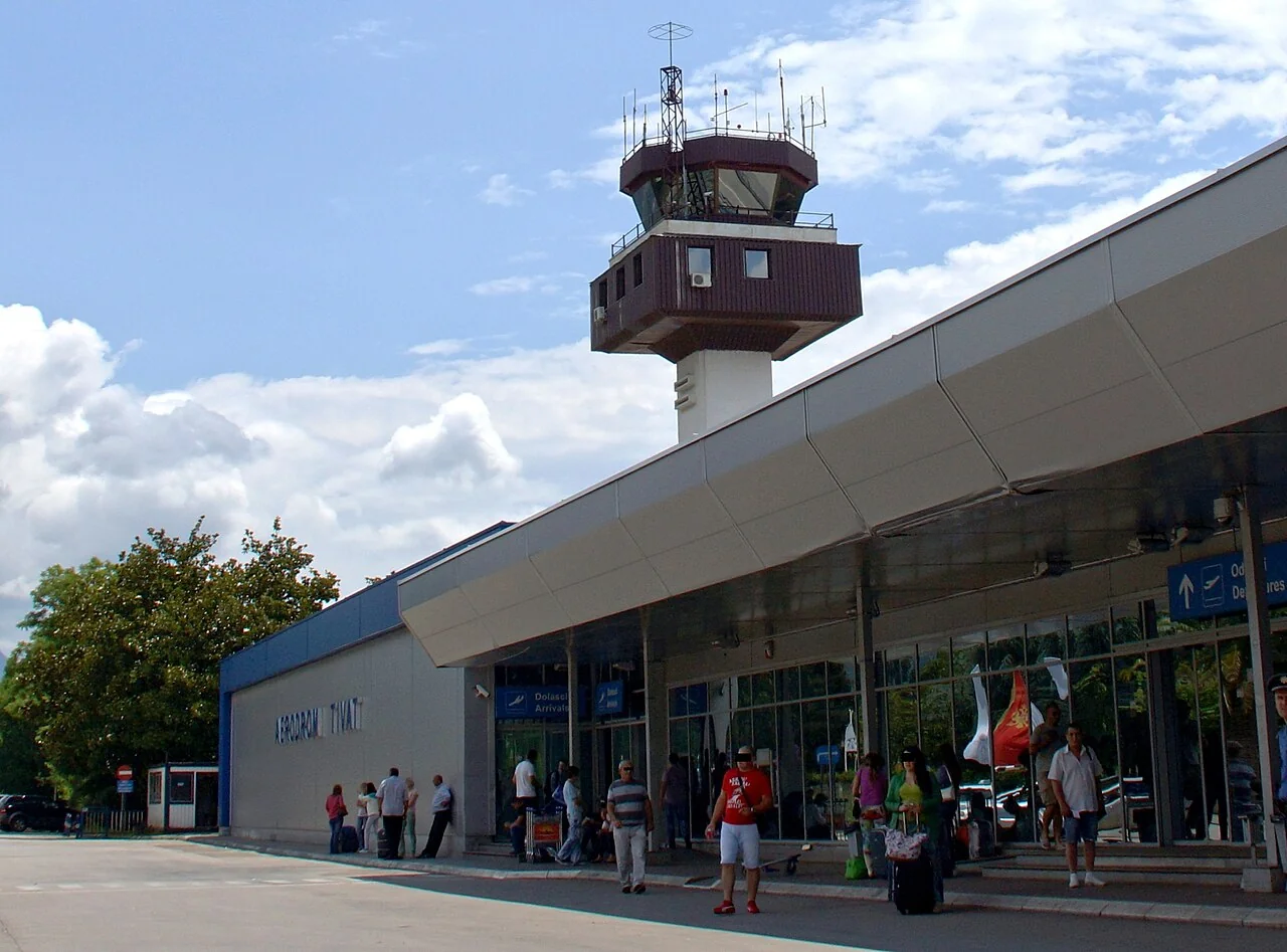 Travellers with luggage outside Tivat Airport terminal, with the control tower behind