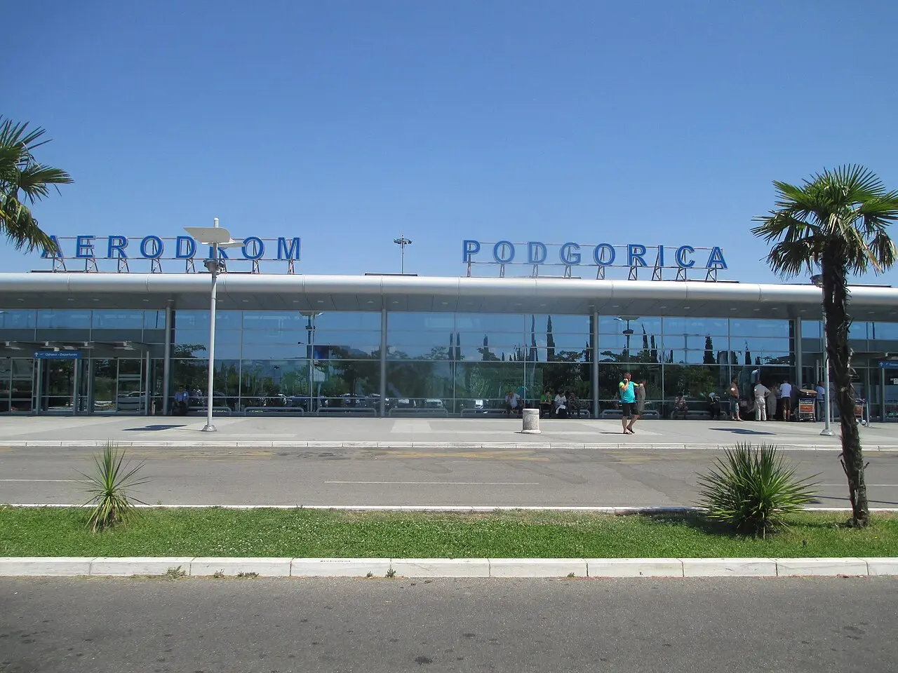 The glass terminal frontage of Podgorica Airport under a clear sky, with the bilingual Aerodrom Podgorica sign