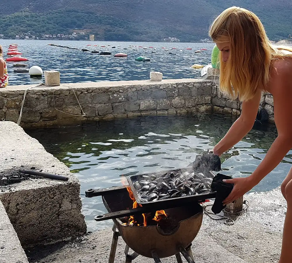 Fresh mussels grilling over a fire at the edge of the Bay of Kotor, with mussel-farm buoys and mountains behind