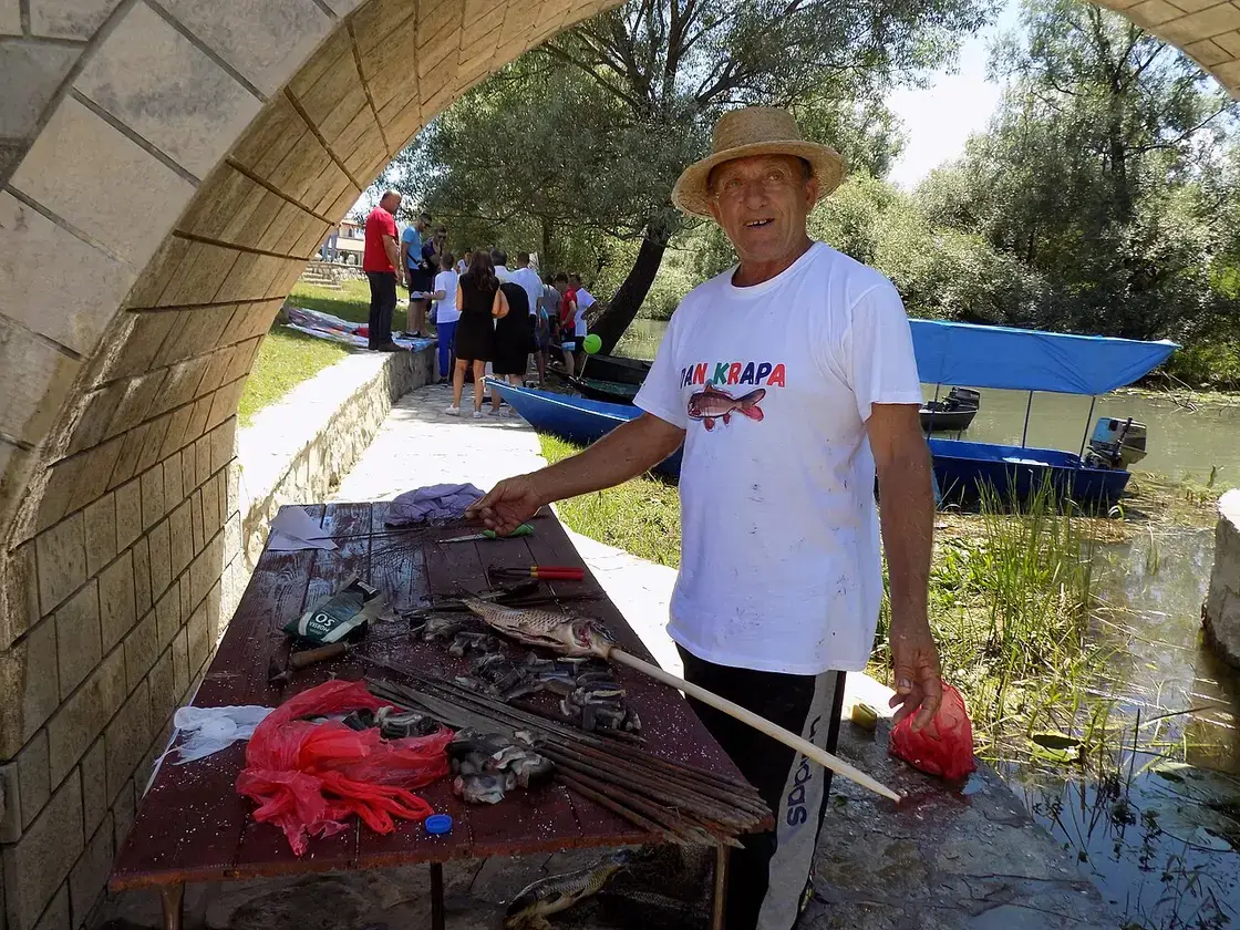 A fisherman preparing carp on skewers by the water at Lake Skadar, with wooden boats behind