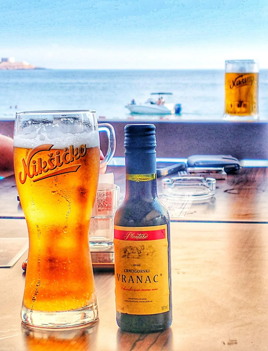 A glass of Nikšićko beer beside a bottle of Plantaže Vranac red wine, with the Adriatic behind