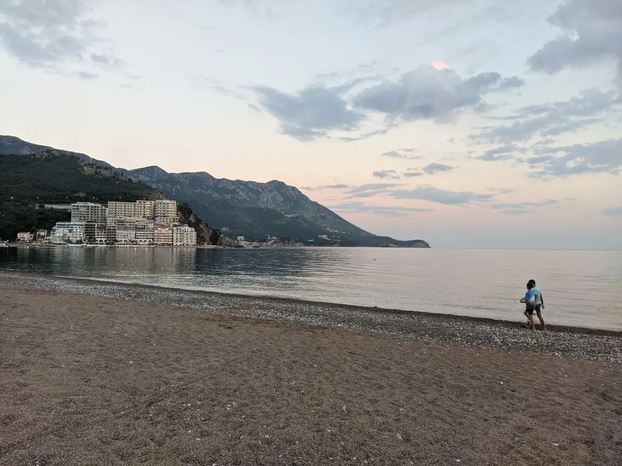 A quiet pebble beach at dusk near Budva with a couple walking and the resort strip lit up along the shore