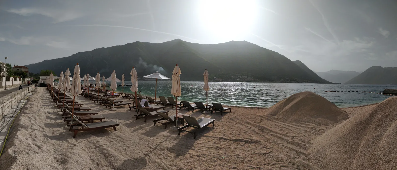 A bay-side beach at Dobrota lined with sun loungers and umbrellas, the calm water of the Bay of Kotor beyond