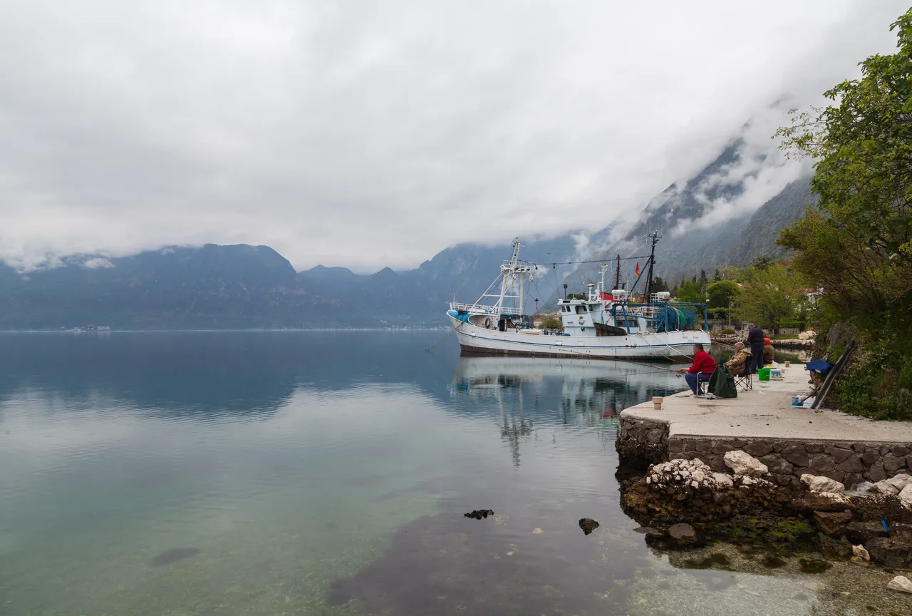 A calm stretch of the Dobrota waterfront on the Bay of Kotor with a moored fishing boat and misty mountains
