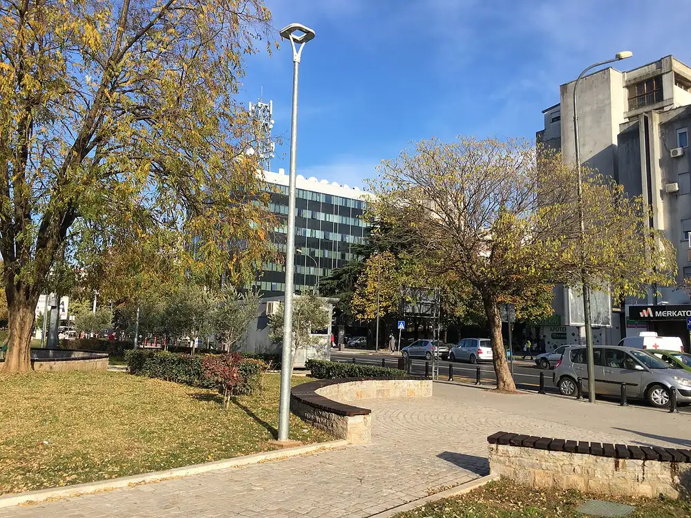 A park with autumn trees and a modern glass office building along a Podgorica boulevard