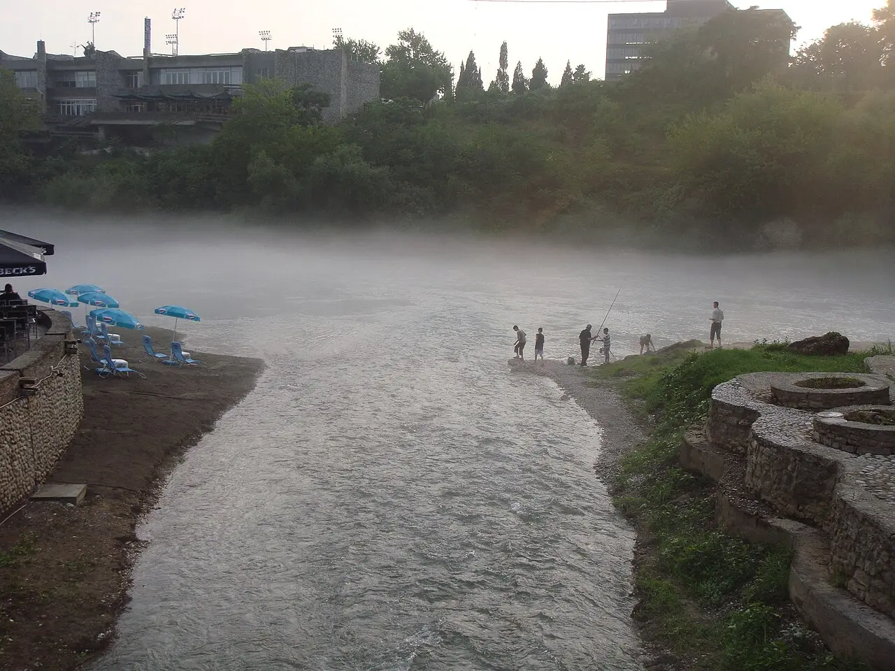 Morning mist rising off the Morača river in Podgorica, with a riverside café and people fishing from the bank