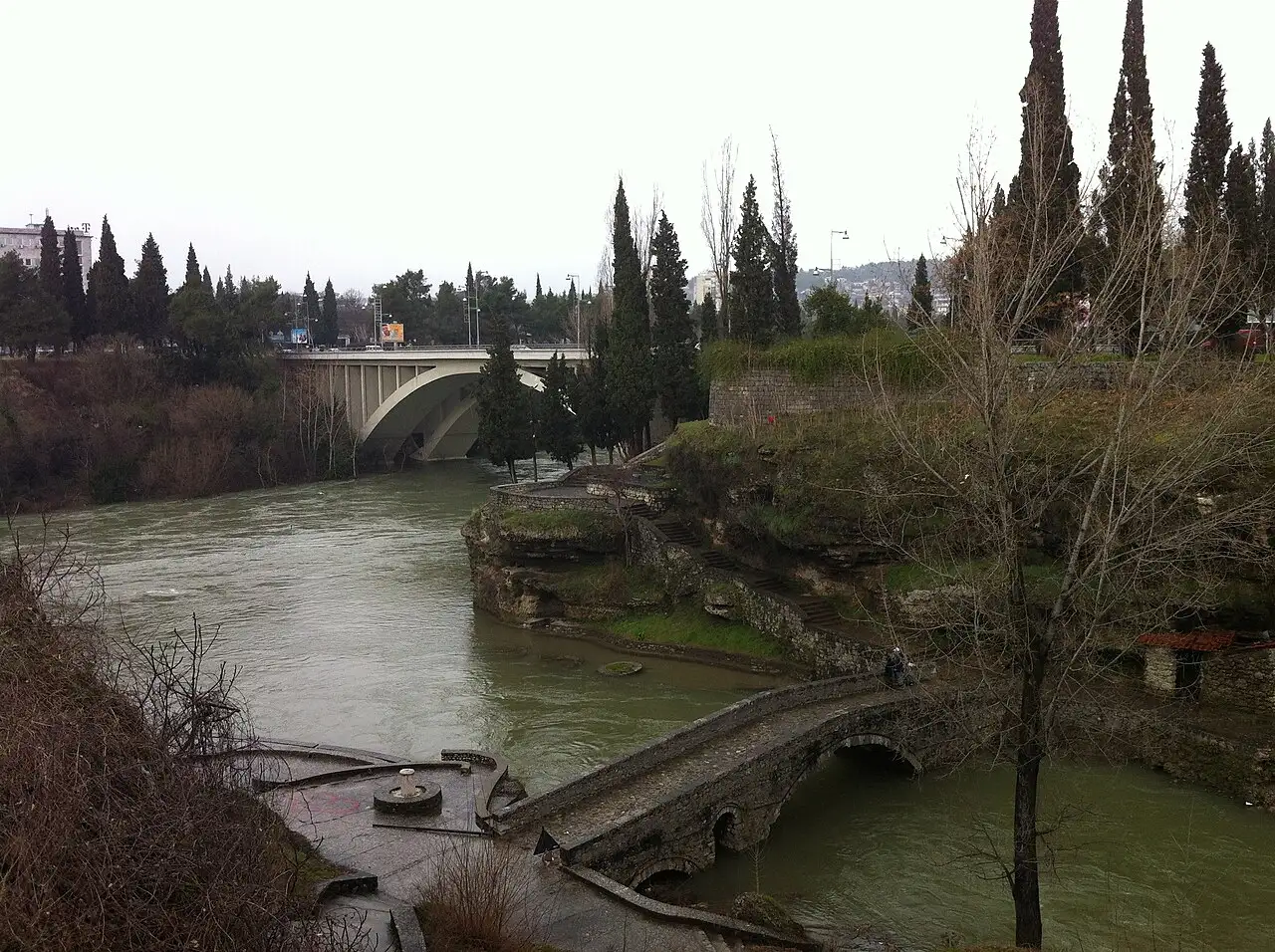 An old stone arch bridge over the Ribnica river near Stara Varoš in Podgorica, with a modern bridge and fortress ruins behind
